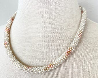 May include: A white pearl necklace with pink and gold accents. The necklace is made of small, round pearls and is strung on a thin wire. The necklace is displayed on a white mannequin.