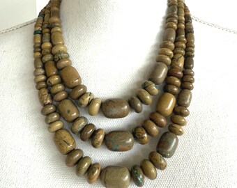 May include: A multi-strand necklace featuring natural-toned, rounded beads in varying sizes. The necklace has a layered design with a mix of smooth and textured beads, showcasing earthy tones of brown and tan. The necklace is displayed on a white mannequin.