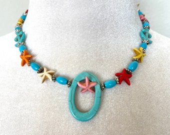 May include: A turquoise blue necklace with a large oval pendant featuring a pink starfish. The necklace is made with turquoise blue, orange, yellow, and white beads and silver accents. The necklace is decorated with starfish charms.