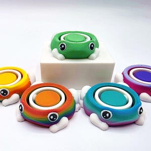 May include: A collection of colorful, frog-shaped, collapsible containers. Each frog has a different color scheme, including green, yellow, orange, blue, and pink. The frogs have white feet and eyes, and are arranged on a white surface.