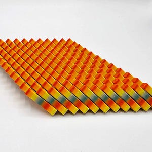 May include: A collection of colorful, geometric wooden blocks arranged in a diagonal pattern. The blocks transition from orange to yellow, with a hint of blue-green in the middle. The arrangement is on a white background.