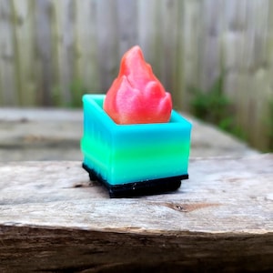 May include: A miniature, colorful flame sculpture. The flame is a vibrant red, set within a square container that transitions from blue to green. The base is black, creating a striking contrast. This decorative piece is ideal for fantasy or gaming enthusiasts.