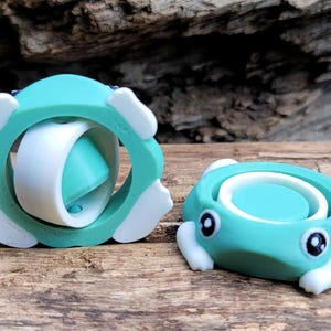 Puede incluir: Dos juguetes de fidget de color verde azulado y blanco. Uno es una esfera con un anillo que gira a su alrededor. El otro es una rana con un anillo giratorio en la espalda.