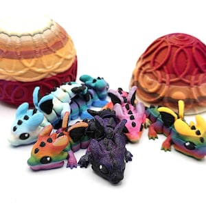 May include: A selection of brightly coloured, 3D-printed dragon figurines in various sizes and hues, including blue, purple, pink, and rainbow. Two decorative, patterned egg-shaped objects are also present, one in shades of red and orange, and the other in yellow and red.