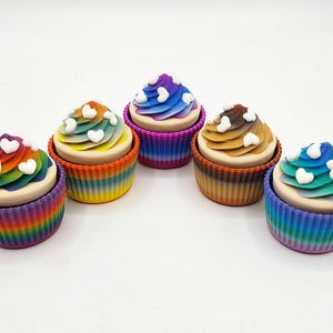 May include: A collection of five colorful cupcake-shaped soaps. Each soap features a rainbow-colored base and a swirl of frosting in various colors, topped with small white heart-shaped decorations. The soaps are arranged on a white surface.