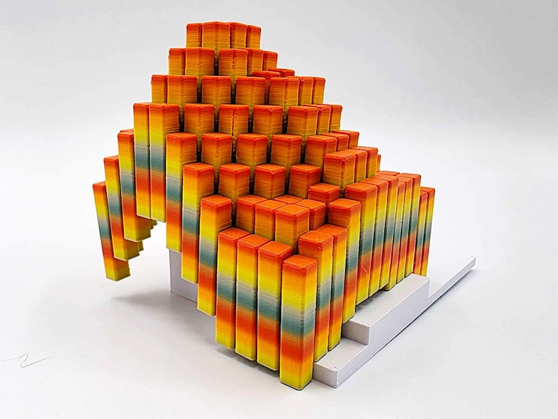 May include: A colorful, abstract sculpture composed of numerous rectangular blocks. The blocks are arranged in a stepped, pyramid-like structure, with a gradient of colors from orange to yellow to green. The base is white.
