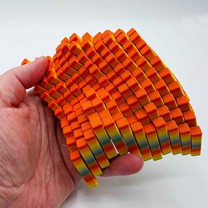 May include: A colorful, abstract sculpture composed of numerous small, rectangular blocks. The blocks transition in color from orange to yellow and blue. The sculpture is held in a hand against a white background.