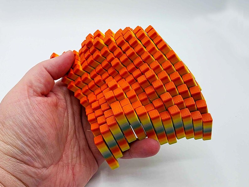 May include: A colorful, abstract sculpture composed of numerous small, rectangular blocks. The blocks transition in color from orange to yellow and blue. The sculpture is held in a hand against a white background.