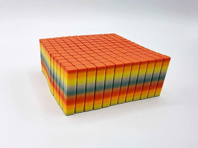 May include: A rectangular block composed of numerous small, colorful, rectangular pieces. The top surface is a solid orange, while the sides reveal a gradient of colors including yellow, green, and orange. The object is set against a white background.