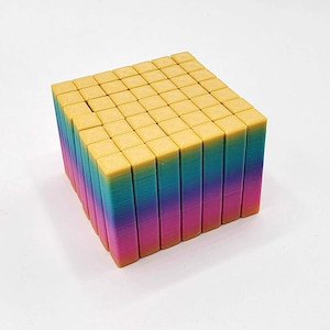 Può includere: Un giocattolo a forma di cubo con la parte superiore gialla e i lati color arcobaleno. Il cubo è composto da molti piccoli blocchi rettangolari. I lati passano dal rosa al blu al giallo. Il giocattolo è probabilmente un puzzle o un set di costruzioni.