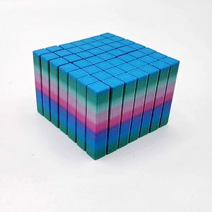 May include: A cube structure made from many small, rectangular blocks. The blocks are arranged to form a larger cube, with a gradient of colours visible on the sides, transitioning from blue to green, pink, and back to blue. The cube is on a white surface.