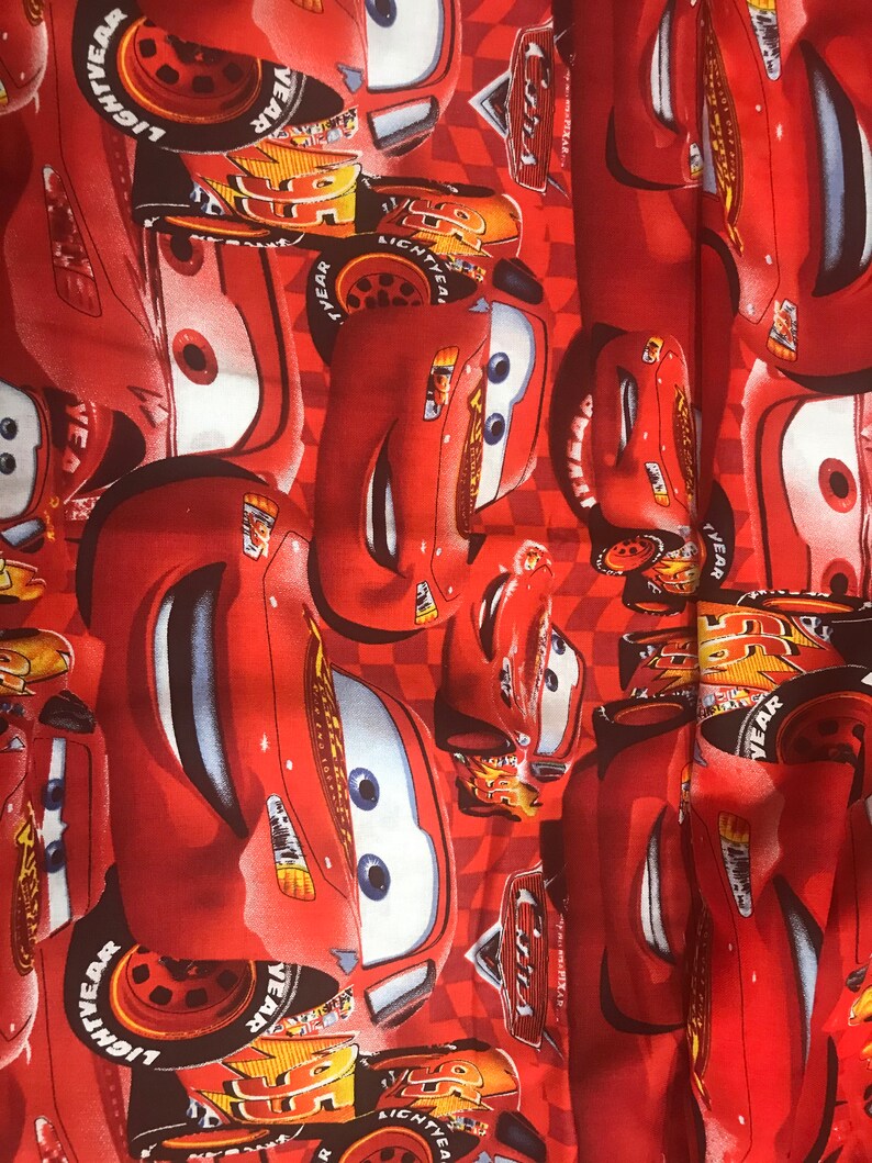 Puede incluir: Tela roja con un patr&oacute;n repetido de coches de carreras rojos de dibujos animados con neum&aacute;ticos negros y las palabras "Lightyear" en los neum&aacute;ticos. Los coches tienen ojos negros y dientes blancos. El fondo es un patr&oacute;n de cuadros rojos y blancos.