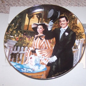 May include: Decorative plate featuring a scene from a classic film. The plate depicts a man in a tuxedo and a woman in a striped dress, with a baby in a basket. The plate has a gold rim and is set against a garden backdrop.