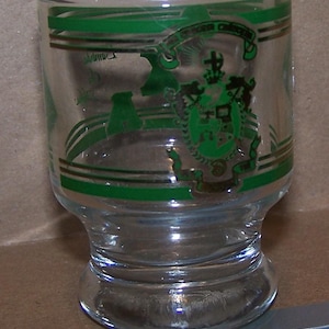 May include: A clear glass tumbler featuring a green and gold printed crest design with a crown and shield motif. The glass has horizontal green and gold bands encircling the body. A silver metal ruler is visible at the base for scale reference.