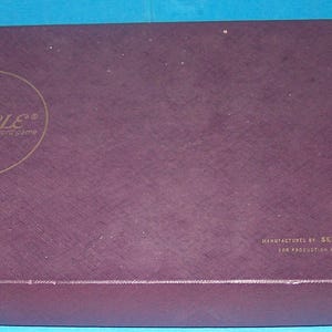 May include: A rectangular, dark purple Scrabble game box. The box has gold lettering and a circular logo on the top. The bottom of the box also has the word "Scrabble" in gold lettering. The box is made in the USA.