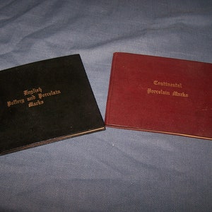 May include: Two hardcover books with gold lettering on the spines. The book on the left is black and says "English Pottery and Porcelain Marks". The book on the right is burgundy and says "Continental Porcelain Marks".
