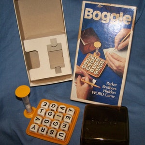 May include: A vintage Boggle word game set. The game includes a box, a dice grid with lettered cubes, a timer, and a game board. The box cover features the game's name and an illustration of hands playing the game.