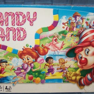 May include: Candy Land board game box with colorful illustrations of characters and a whimsical landscape. The game title is in pink candy-cane style letters. The box is labeled "Preschool" and "Ages 3+".