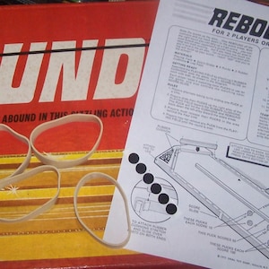 May include: A vintage board game box with the word "REBOUND" in large white letters. The box is red and yellow, with several rubber bands and a game instruction sheet.
