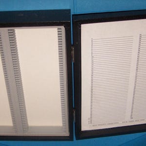 May include: A black wooden box with two compartments. One side has vertical dividers, the other has a white sheet with numbered lines. The box is open, revealing the interior.