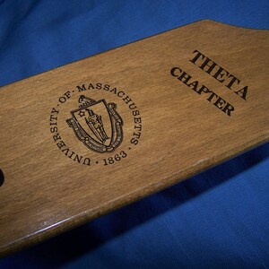 May include: A wooden paddle with the text "Theta Chapter" and "University of Massachusetts 1863" engraved on it. The paddle also features a seal with the state of Massachusetts coat of arms.