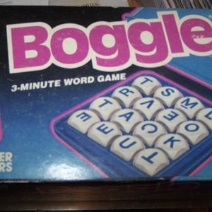 May include: A blue and white Boggle game box with a pink title and a 3-minute word game label. The box includes a blue and white letter cube tray and an hourglass.