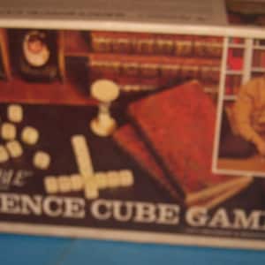 May include: A vintage Scrabble Sentence Cube Game box with a red, white, and green cube container, white dice, and a brown and white book. The box has a picture of two people playing the game.