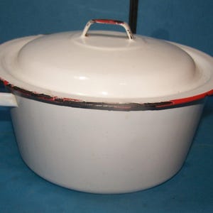 May include: White enamelware pot with a lid. The pot has a rounded shape with a lid and two side handles. The rim and handles are accented with red. The pot shows signs of wear.