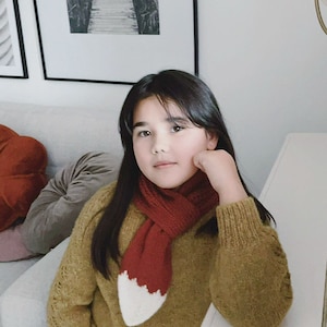 May include: A young person wearing a rust-colored scarf with a white fox tail design, paired with a mustard-yellow sweater. The scarf is knitted and adds a playful touch to the outfit. The Instagram handle @annaluciadu is visible in the image.