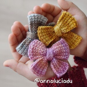 May include: Three crocheted bows in different colors: light blue, yellow, and pink. The bows are held in a hand.
