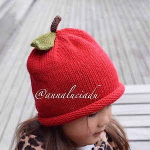 Könnte beinhalten: Eine rote Strickmütze in Form eines Apfels mit einem grünen Filzblatt oben. Die Mütze wird von einem Kind mit langen braunen Haaren und einer Jacke mit Leopardenmuster getragen. Der Text "@annaluciadu" ist auf der Mütze sichtbar.