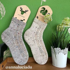 Könnte beinhalten: Zwei Paar weiße gestrickte Socken mit einem Spitzenmuster. Die Socken werden auf Holzsockensperren mit Tier-Ausschnitten präsentiert. Der Text "Annalucia Design" ist auf den Sockensperren sichtbar.