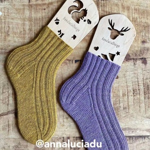 Può includere: Due paia di calze lavorate a mano esposte su blocca-calze in legno. Le calze sono a coste e presentano un colore giallo senape e un colore viola. I blocca-calze sono decorati con uno scoiattolo e un cervo, e il testo "Annalucia Design" è stampato su ogni blocca-calze.