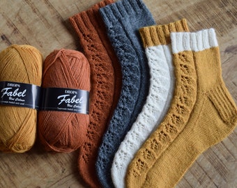 May include: Four pairs of knitted socks in different colors: mustard yellow, orange, white, and gray. The socks have a lacy pattern and are made with Drops Fabel yarn.