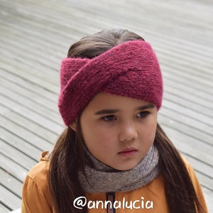 May include: A young person wearing a burgundy knitted headband with a criss-cross design. The headband is worn over long brown hair. The person is wearing a yellow jacket and a grey scarf. The background is a wooden deck.