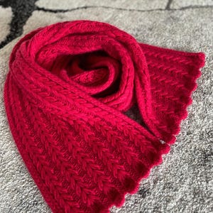 May include: A bright red knitted scarf with a textured pattern and scalloped edges. The scarf is made of a soft, thick yarn and is draped in a spiral shape. This cosy accessory is perfect for cold weather.