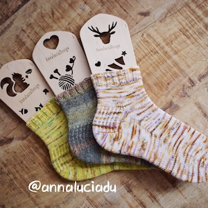 Puede incluir: Tres pares de calcetines de punto a mano coloridos que se muestran en bloqueadores de calcetines de madera con recortes de un ciervo, una ardilla y una bola de lana. Los bloqueadores de calcetines están etiquetados como "Annalucia Design". Los calcetines se muestran sobre una superficie de madera. La imagen incluye el texto "@annaluciadu".