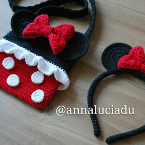 Pu&ograve; includere: Una borsa a mano in crochet rosso e nero con pois bianchi e un volant in crochet bianco. La borsa ha una tracolla in crochet nero e un fiocco in crochet rosso. Un cerchietto in crochet nero con orecchie in crochet rosso e un fiocco in crochet rosso. Il cerchietto ha il testo "@annaluciadu" sulla superficie in legno.