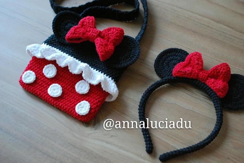 Pu&ograve; includere: Una borsa a mano in crochet rosso e nero con pois bianchi e un volant in crochet bianco. La borsa ha una tracolla in crochet nero e un fiocco in crochet rosso. Un cerchietto in crochet nero con orecchie in crochet rosso e un fiocco in crochet rosso. Il cerchietto ha il testo "@annaluciadu" sulla superficie in legno.