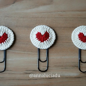 May include: Three handmade crochet heart bookmarks. Each bookmark features a white circular design with a red heart in the center, attached to a black paperclip. The bookmarks are arranged on a wooden surface. The text "@annaluciadu" is visible.