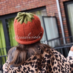 May include: An orange knitted hat with a green leaf design. The hat is worn by a person wearing a leopard print jacket. The text "@annaluciadu" is visible on the hat.