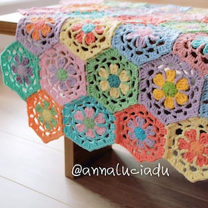 May include: A colorful, handmade crochet blanket with a floral design. The blanket is made of hexagonal pieces in pastel colors, including pink, yellow, blue, and green. The flowers in the center of each hexagon are in contrasting colors. The blanket is draped over a wooden table.