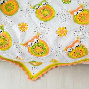 Könnte beinhalten: Eine weiße gehäkelte Babydecke mit einem gewellten Rand. Die Decke zeigt ein sich wiederholendes Muster aus Eulen und Blumen in Orange-, Grün- und Gelbtönen.