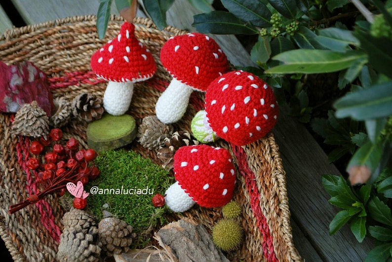 May include: Four crocheted red and white mushrooms with white stems are arranged in a woven basket with other natural elements, including pine cones, moss, and berries.