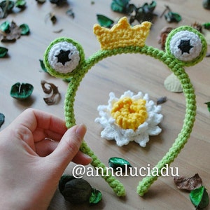 Può includere: Fascia per capelli in crochet verde con orecchie di rana e una corona gialla. La fascia ha occhi neri con pupille bianche. La fascia è su una superficie di legno con foglie e fiori verdi.