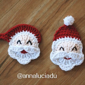 May include: Two crocheted Santa Claus faces. One Santa has a red hat with a white pom-pom, a white beard, and a smiling face. The other Santa has a red hat with a white brim and a white beard. The faces are made of cream, red, and white yarn.