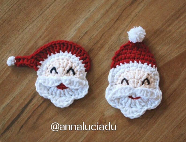 May include: Two crocheted Santa Claus faces. One Santa has a red hat with a white pom-pom, a white beard, and a smiling face. The other Santa has a red hat with a white brim and a white beard. The faces are made of cream, red, and white yarn.