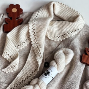 以下が含まれることがあります： ベージュのカーディガンに、スカラップエッジのクリーム色のニットショールがかけられています。クリーム色の糸玉と、木製の花の形をした装飾品も見えます。「Andalucia Designs」と書かれた糸のラベル。