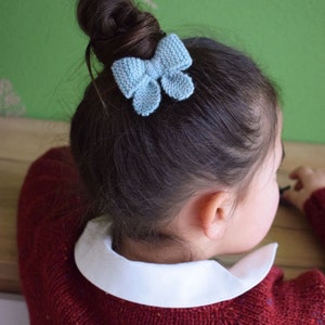 May include: A young girl with brown hair in a bun, wearing a red jumper with a white collar, and a light blue knitted bow in her hair. The background is a green wall with a white pattern.
