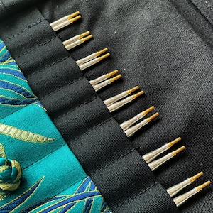 Può includere: Primo piano di un set di ferri da maglia con punte in metallo argentato e dettagli dorati, ordinatamente disposti in un astuccio di tessuto nero. L'astuccio è parzialmente aperto, rivelando i ferri e un tessuto fantasia turchese e oro.